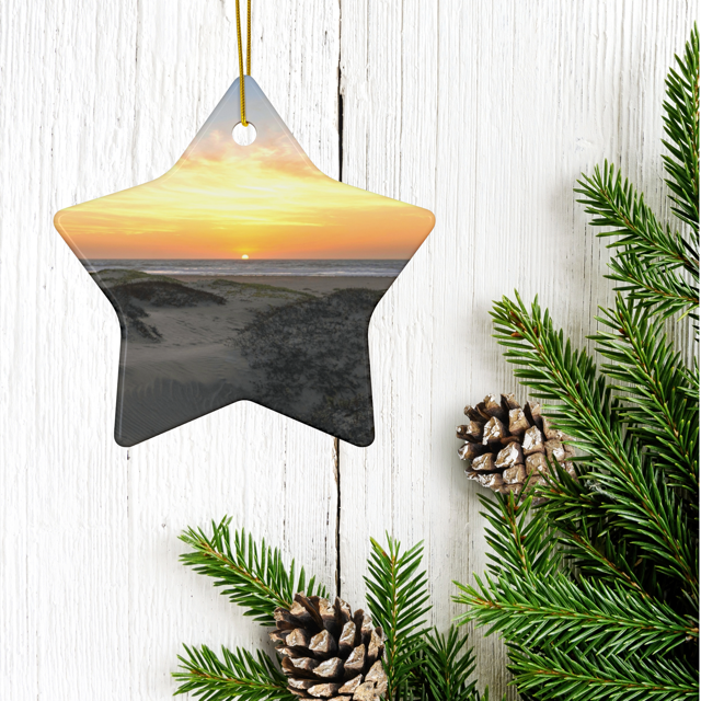 Ceramic Christmas ornament featuring an original photograph of a sunset over coastal sand dunes on a wooden surface with pine branches and cones.