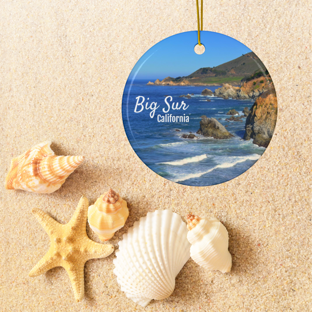Ceramic Christmas ornament featuring a photograph of the rugged cliffs and deep blue Pacific Ocean along the Big Sur coastline in California, surrounded by seashells on sand.