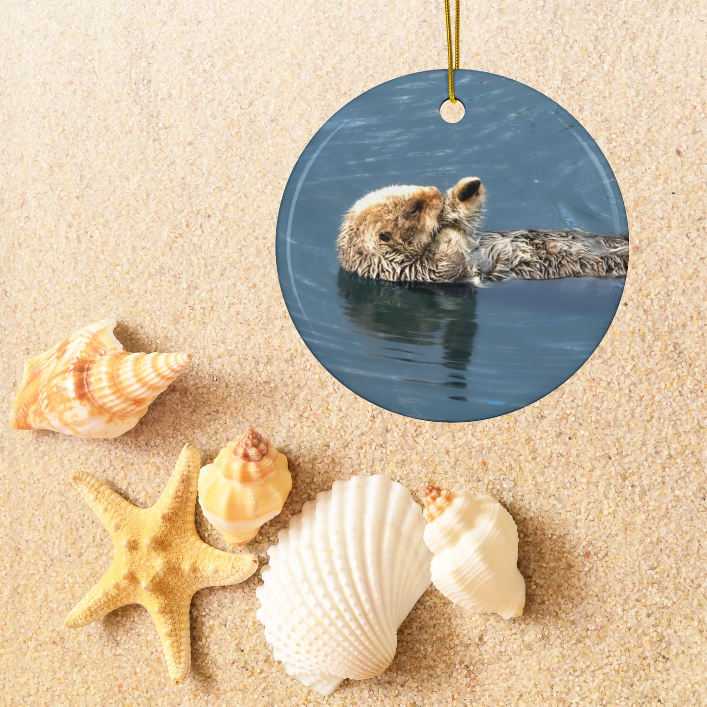 Round ornament featuring a professional photograph of a sea otter floating on its back in the blue waters of the Pacific Ocean on sand with shells and starfish