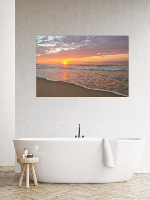 Canvas print of a vibrant sunrise over the Atlantic Ocean on a wall above a white bathtub in a bathroom.