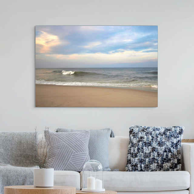 A canvas print of a peaceful view of tiered ocean waves rolling on to a sandy beach under a soft peach and blue cloud-filled sky on a wall above a sofa with decorative pillows.