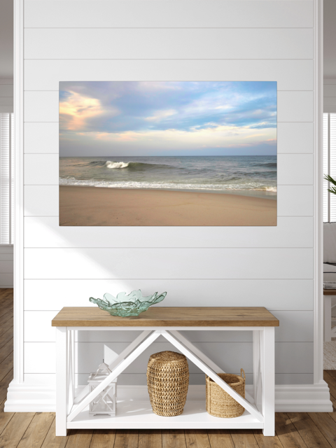 A canvas print of a peaceful view of tiered ocean waves rolling on to a sandy beach under a soft peach and blue cloud-filled sky on a wall above a wooden bench with shelves.