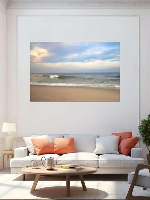 Living room with a large canvas print of a peaceful view of tiered ocean waves rolling on to a sandy beach under a soft peach and blue cloud-filled sky on the wall above a white sofa with orange and white cushions.
