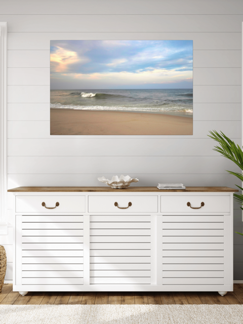 White cabinet with wooden top against a wall with a canvas print of a peaceful view of tiered ocean waves rolling on to a sandy beach under a soft peach and blue cloud-filled sky