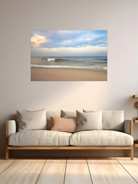 A canvas print of a peaceful view of tiered ocean waves rolling on to a sandy beach under a soft peach and blue cloud-filled sky on a wall above a sofa with cushions.