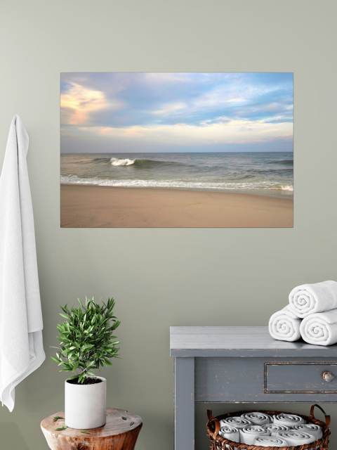 A canvas print of a peaceful view of tiered ocean waves rolling on to a sandy beach under a soft peach and blue cloud-filled sky on a wall above a plant and shelves with towels.