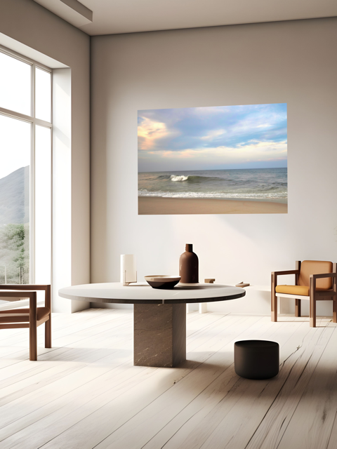 Modern living room with a large canvas print of a peaceful view of tiered ocean waves rolling on to a sandy beach under a soft peach and blue cloud-filled sky, wooden chairs, and a round table.