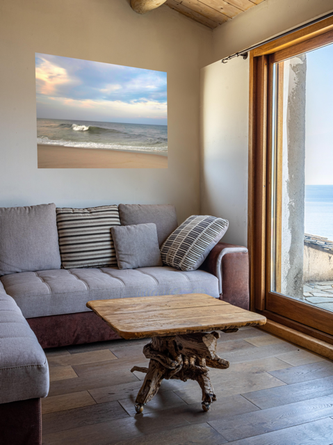 Living room with a gray sofa, wooden coffee table, and a canvas print of a peaceful view of tiered ocean waves rolling on to a sandy beach under a soft peach and blue cloud-filled sky.