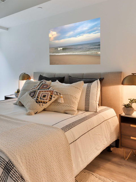 Bedroom with a bed featuring decorative pillows and a large canvas print of a peaceful view of tiered ocean waves rolling on to a sandy beach under a soft peach and blue cloud-filled sky.