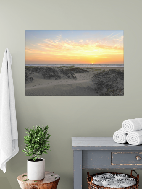 A canvas print of a beach dunes sunset on a wall above a table with a plant and folded towels.