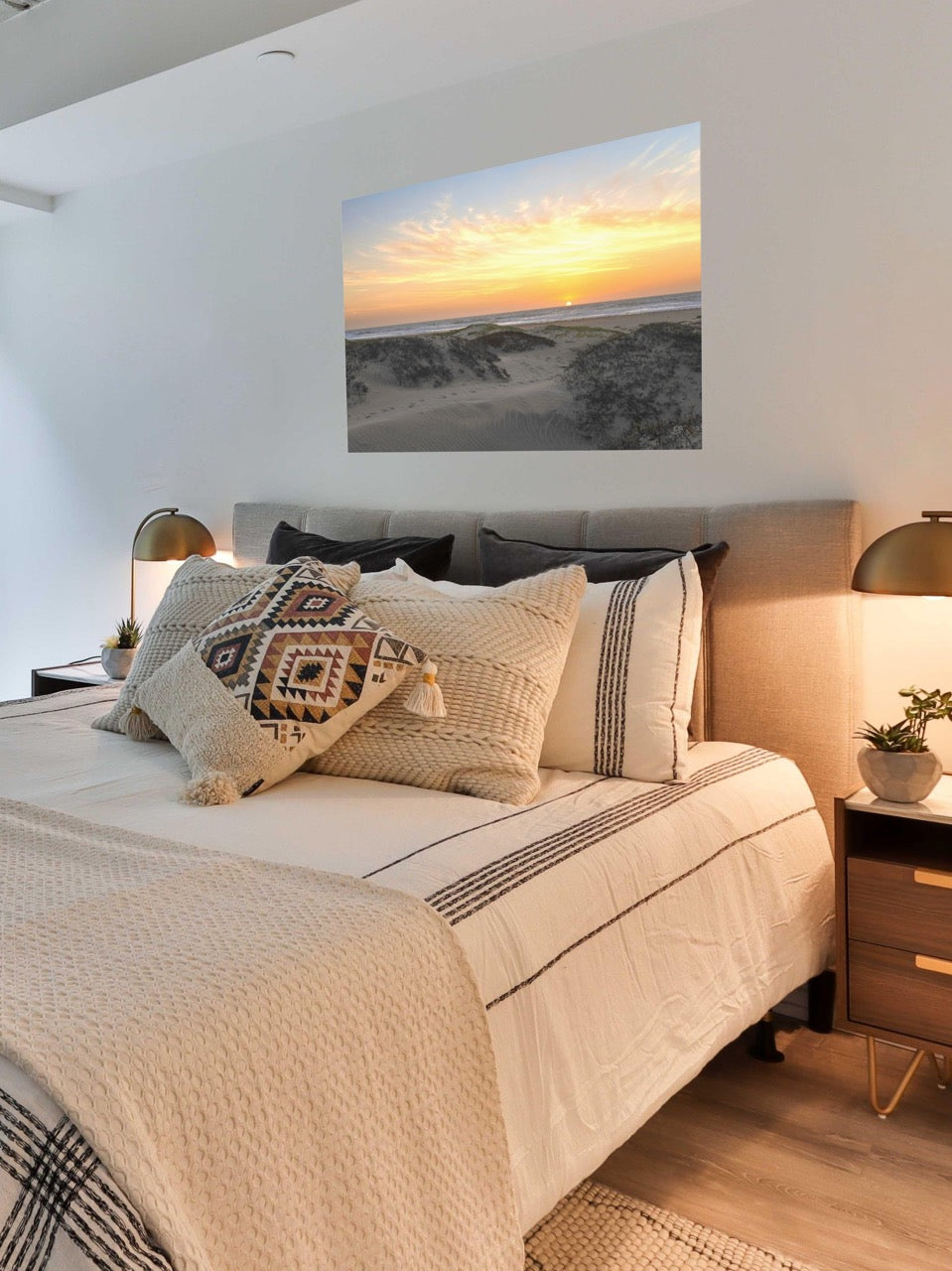 Bedroom with a bed featuring patterned pillows, a canvas print of a beach dunes sunset on the wall, and nightstands with lamps.