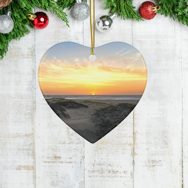 Heart-shaped ceramic Christmas ornament featuring an original photograph of a sunset over coastal sand dunes on a wooden surface with festive decorations.