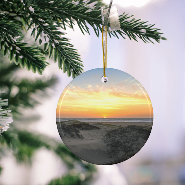 Ceramic Christmas ornament featuring an original photograph of a sunset over coastal sand dunes hanging on a Christmas tree branch.
