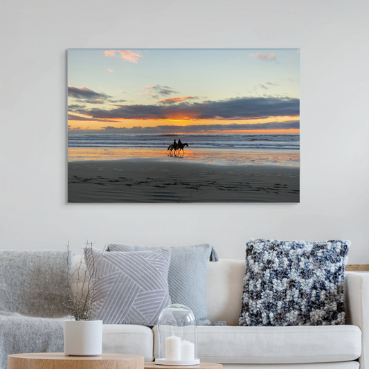 A canvas print of two horseback riders on the beach at sunset on a white wall above a sofa.