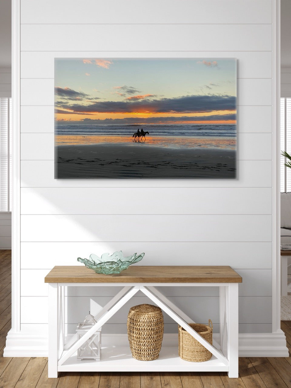 A canvas print of two horseback riders on the beach at sunset on a wall above a wooden bench.
