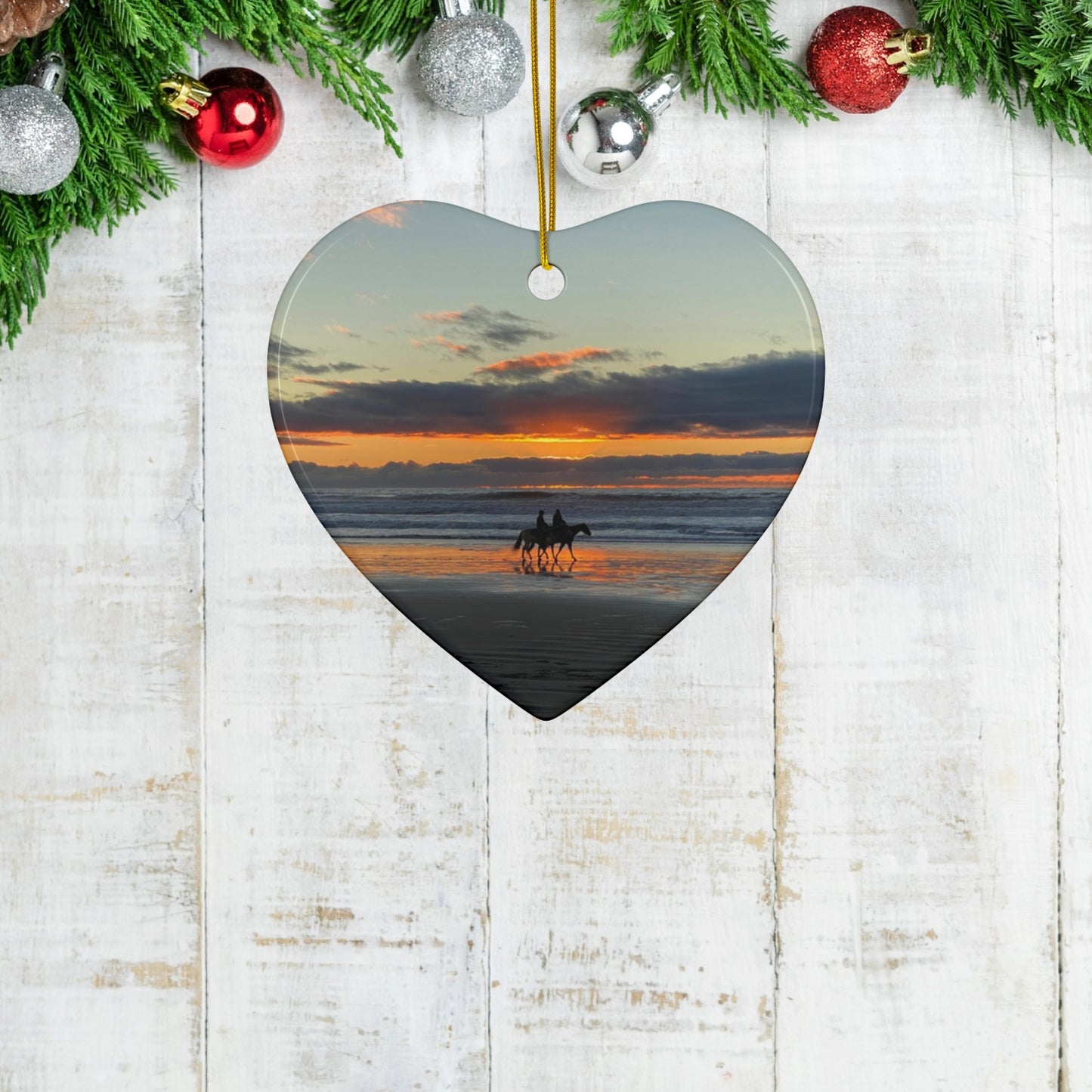 Heart-shaped ornament with a sunset beach scene and two horses, hanging on a wooden surface with Christmas decorations.