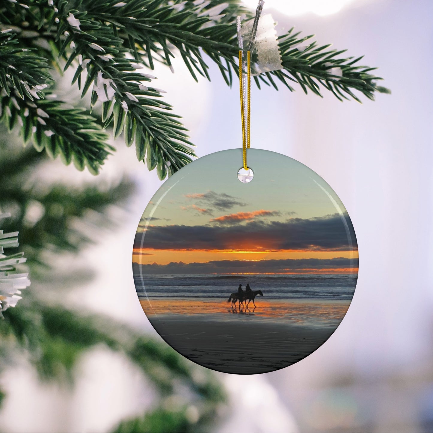 Ceramic holiday ornament featuring a photograph of two horseback riders silhouetted on a beach at sunset with reflections in the wet sand, hanging on a Christmas tree.