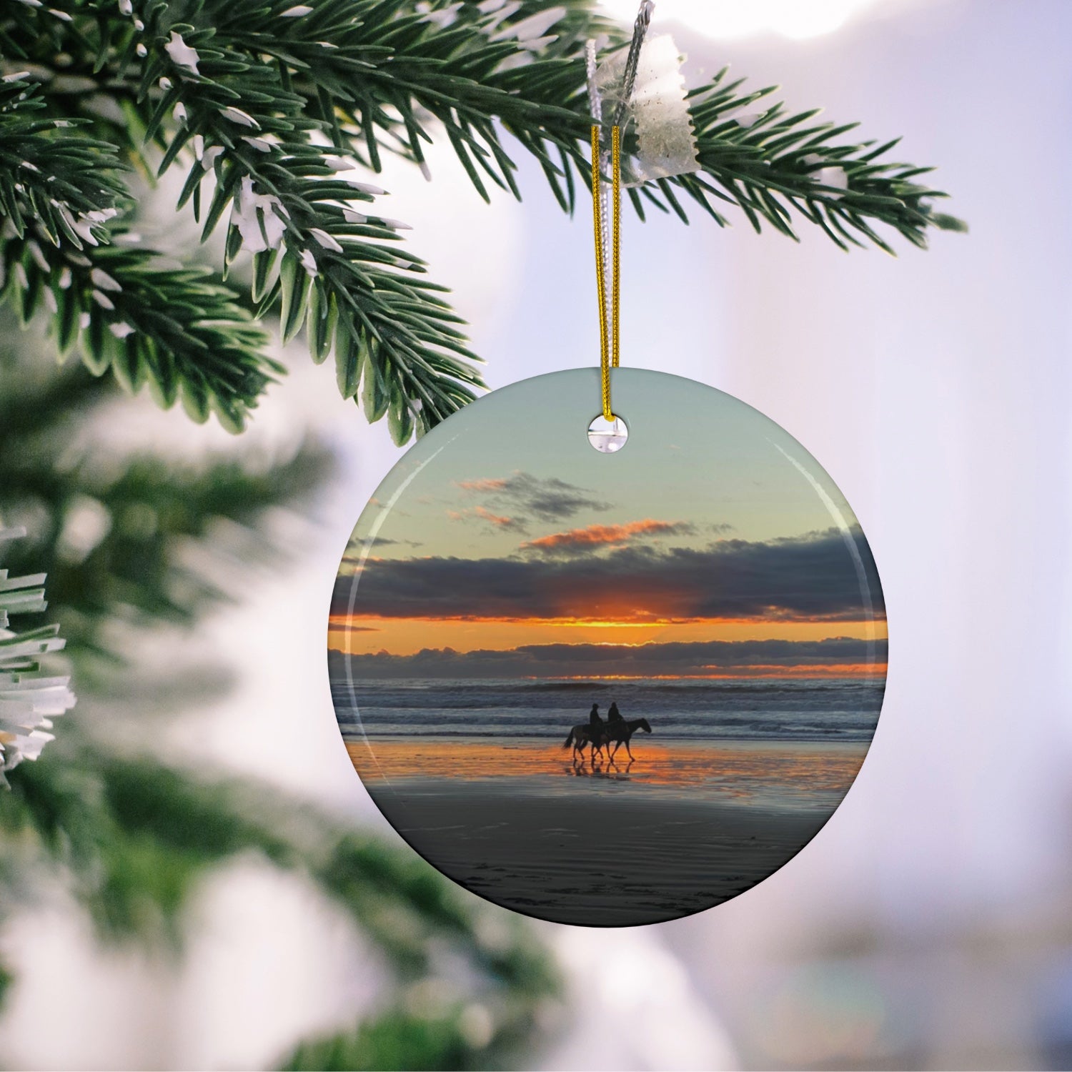 Ceramic holiday ornament featuring a photograph of two horseback riders silhouetted on a beach at sunset with reflections in the wet sand, hanging on a Christmas tree.