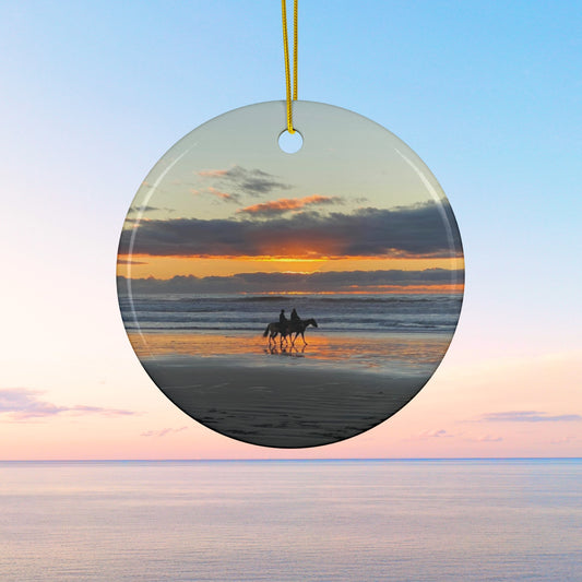 Ceramic holiday ornament featuring a photograph of two horseback riders silhouetted on a beach at sunset with reflections in the wet sand.