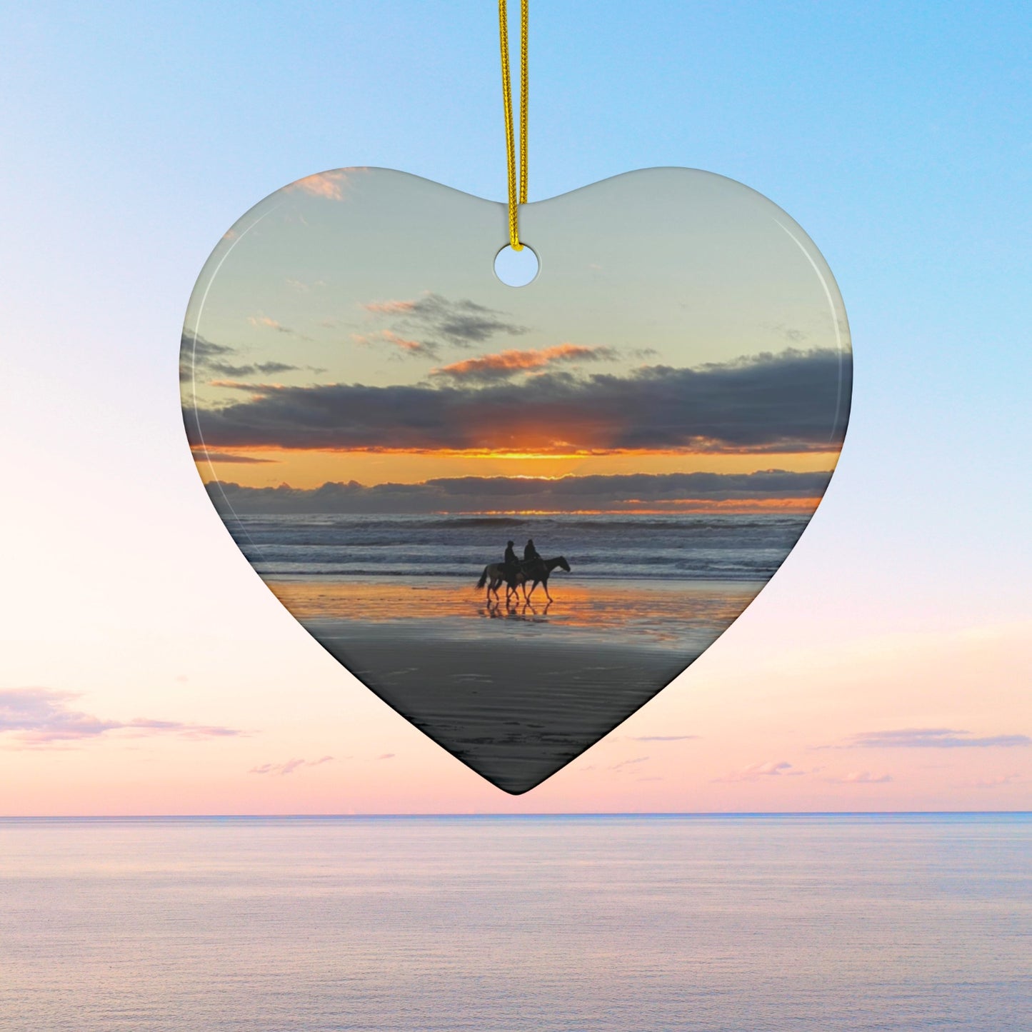 Heart-shaped ceramic holiday ornament featuring a photograph of two horseback riders silhouetted on a beach at sunset with reflections in the wet sand.