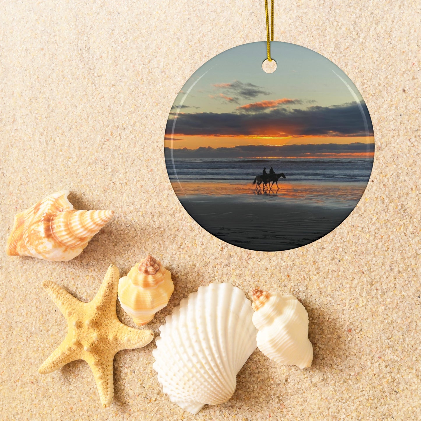 Ceramic holiday ornament featuring a photograph of two horseback riders silhouetted on a beach at sunset with reflections in the wet sand, surrounded by seashells on sand.