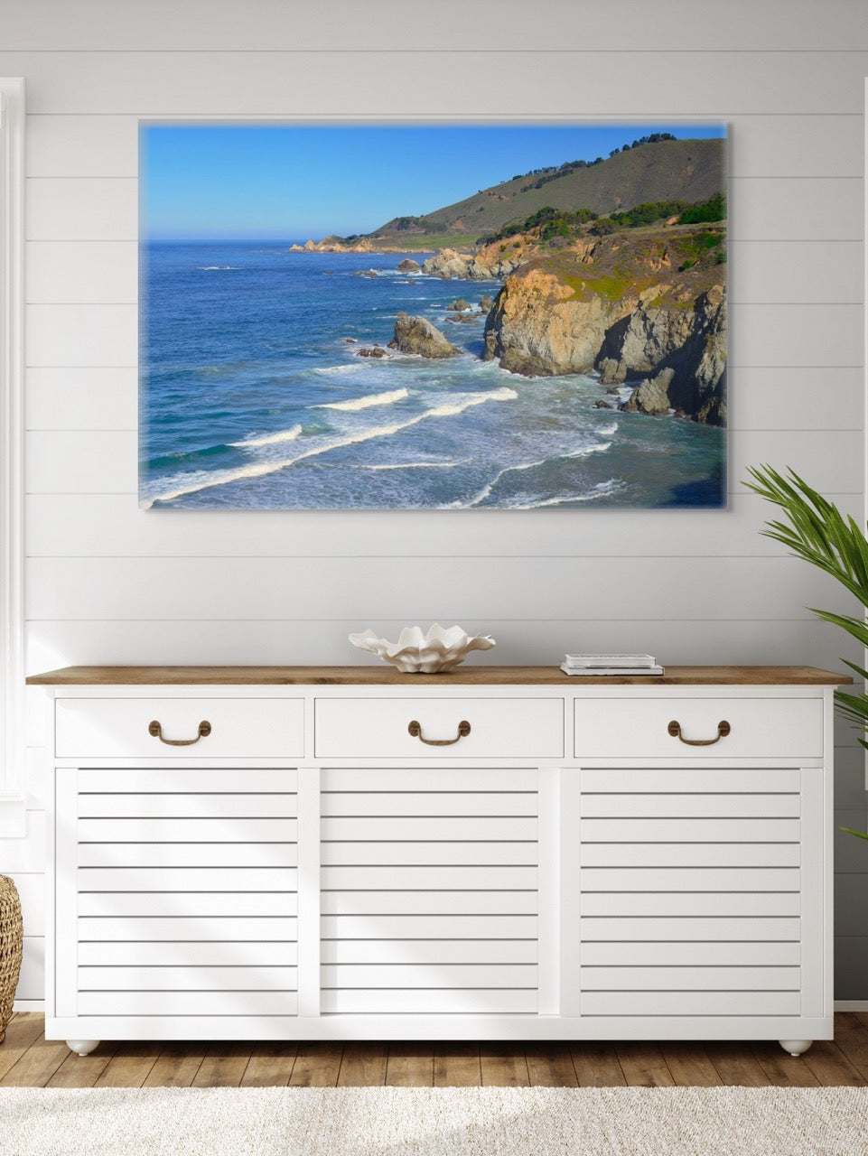 White cabinet with wooden top below a large canvas print of the rugged coastline in Big Sur, California on a white wall.