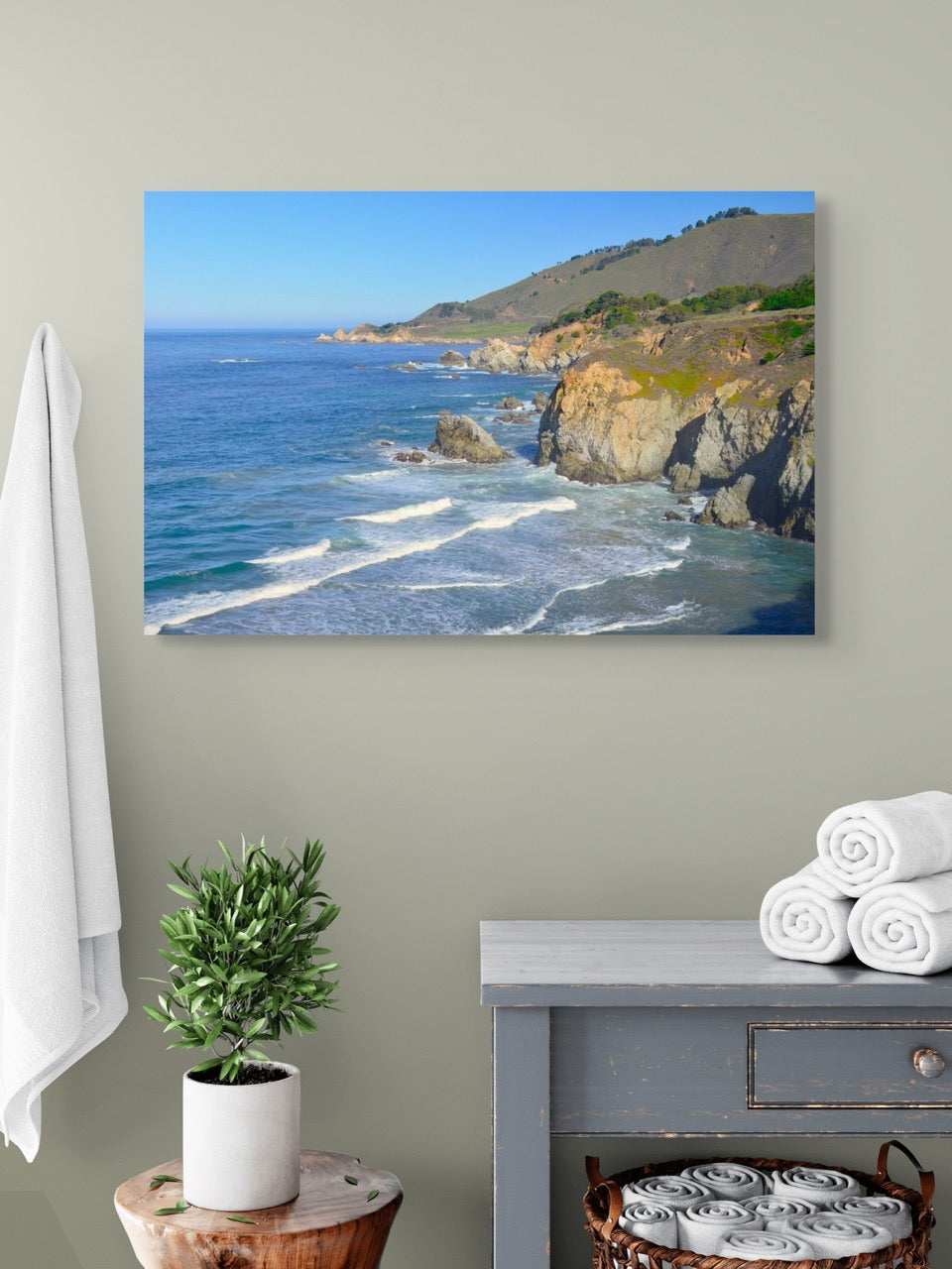 A canvas print of the rugged coastline in Big Sur, California hanging on a wall above a dresser with towels and a plant.