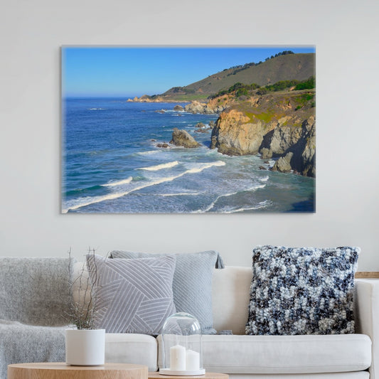 A canvas print of the rugged coastline in Big Sur, California on a wall above a living room sofa with decorative pillows.