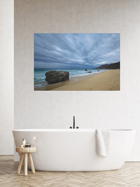 Canvas Print of a Big Sur beach with a stormy sky on a wall above a white bathtub in a bathroom.