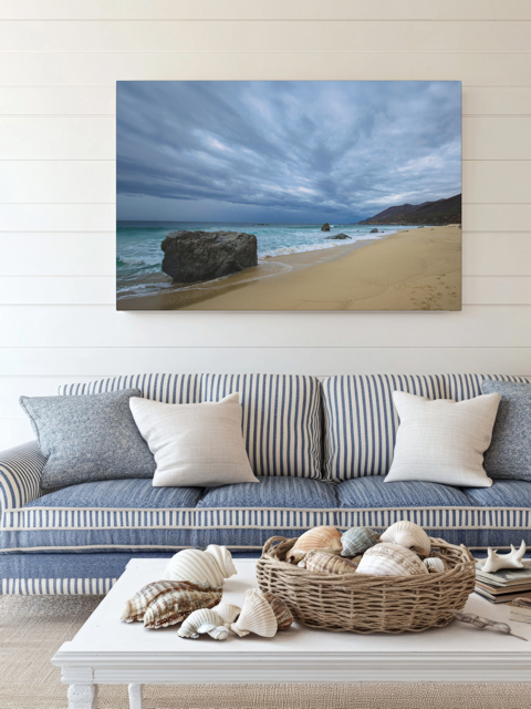 Canvas print of a Big Sur beach with a stormy sky above a striped sofa with decorative items on a coffee table.
