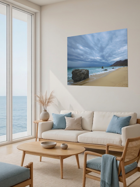 Living room with a large window view of the ocean, a beige sofa with blue cushions, and a canvas print of a Big Sur beach with a stormy sky on the wall.