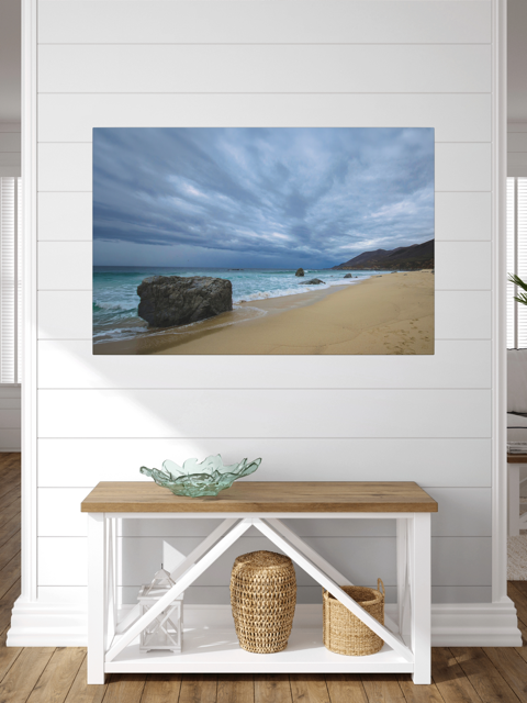 Canvas print of a Big Sur beach with a stormy sky on a wall above a wooden bench with white shelves.
