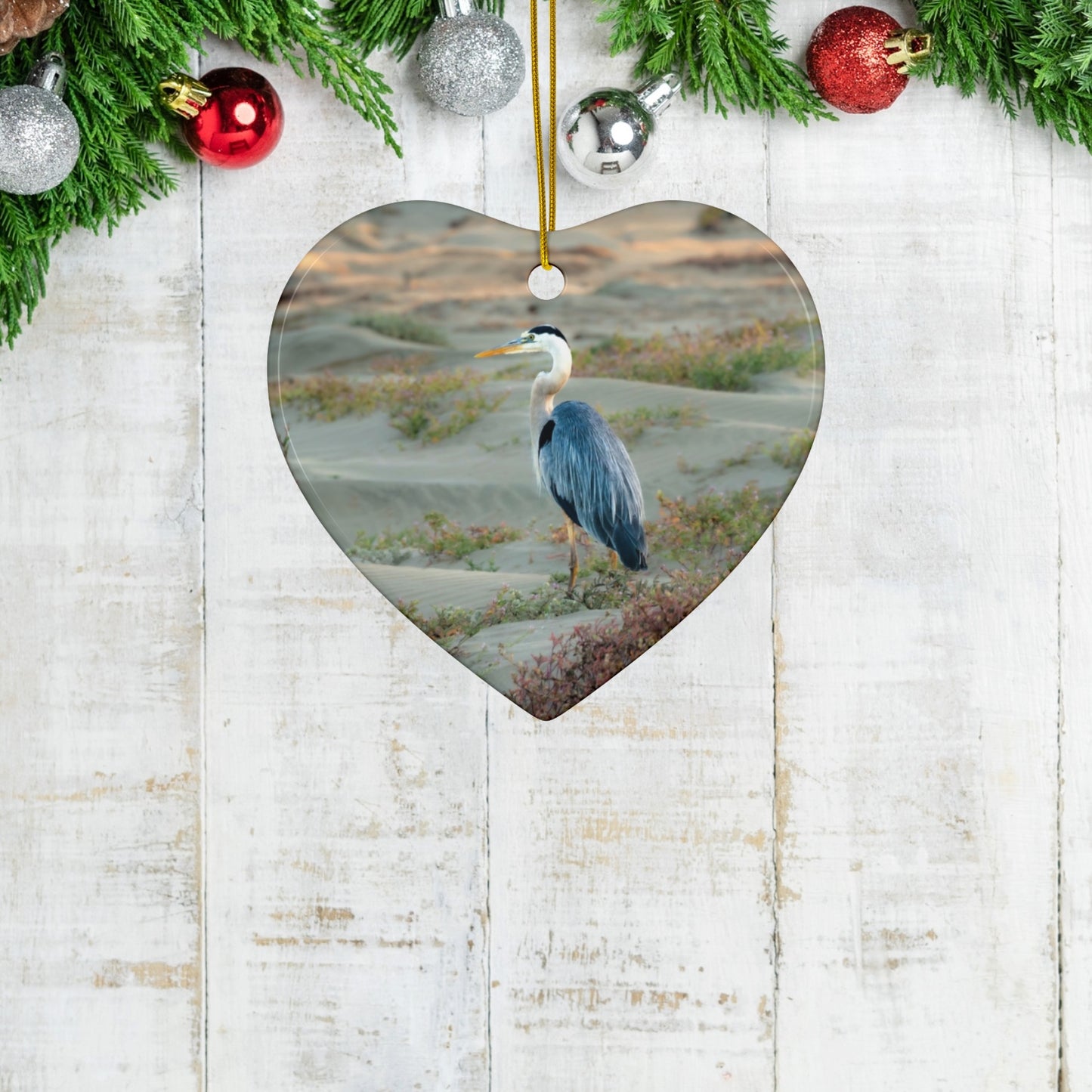 Heart-shaped ceramic holiday ornament featuring a professional photograph of a Great Blue Heron standing among coastal reeds along the Pacific shoreline on a wooden background with Christmas decorations