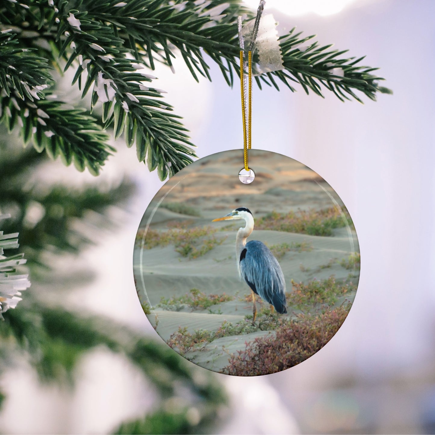 Ceramic holiday ornament featuring a professional photograph of a Great Blue Heron standing among coastal reeds along the Pacific shoreline hanging on a tree branch.
