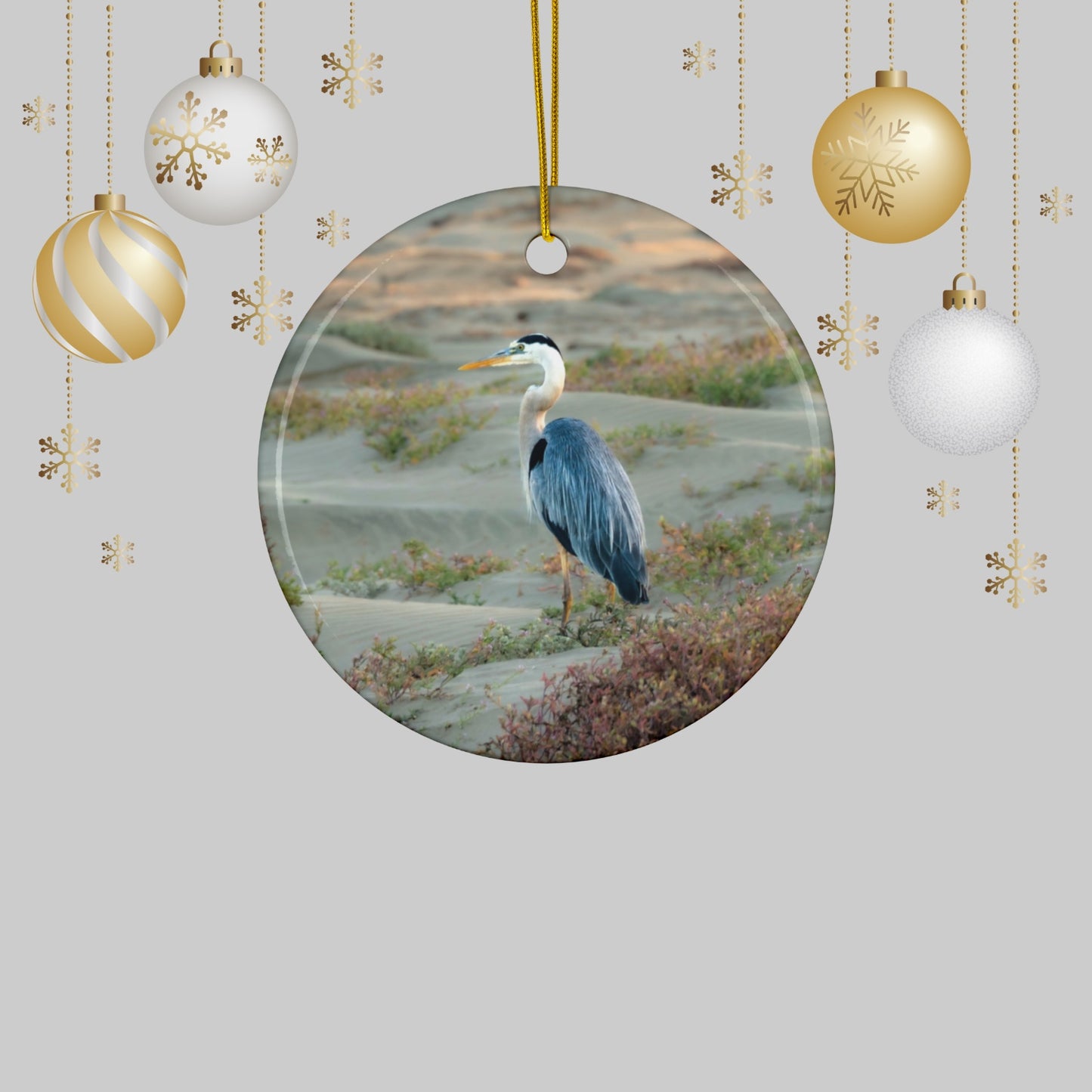 Ceramic holiday ornament featuring a professional photograph of a Great Blue Heron standing among coastal reeds along the Pacific shoreline, surrounded by Christmas ornaments and decorations.