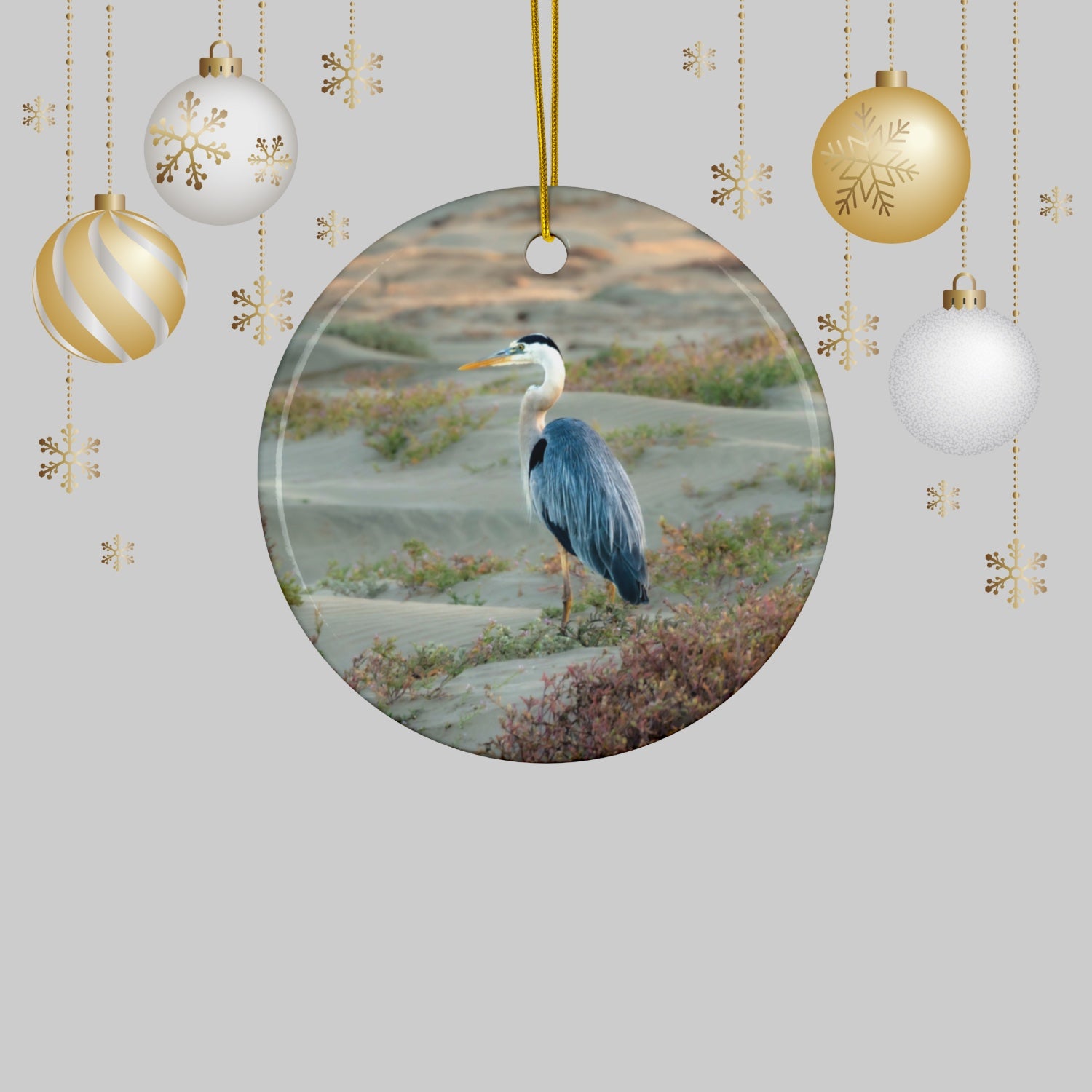 Ceramic holiday ornament featuring a professional photograph of a Great Blue Heron standing among coastal reeds along the Pacific shoreline, surrounded by Christmas ornaments and decorations.