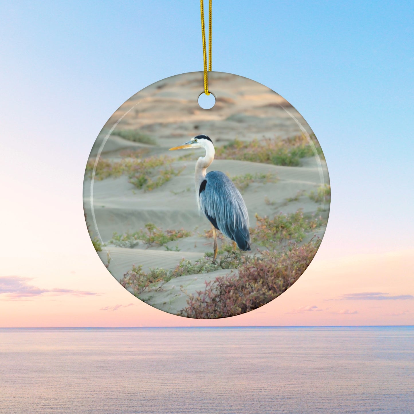 Ceramic holiday ornament featuring a professional photograph of a Great Blue Heron standing among coastal reeds along the Pacific shoreline.