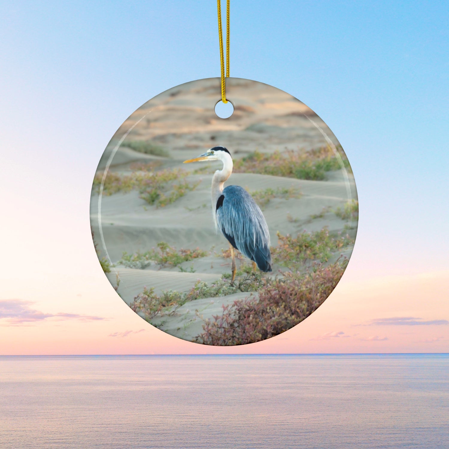 Ceramic holiday ornament featuring a professional photograph of a Great Blue Heron standing among coastal reeds along the Pacific shoreline.