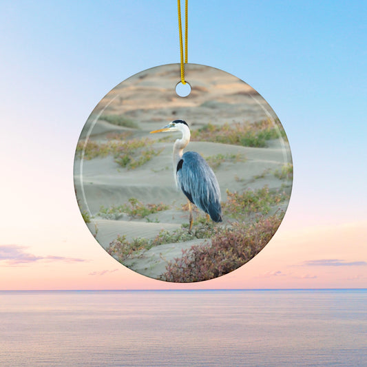Ceramic holiday ornament featuring a professional photograph of a Great Blue Heron standing among coastal reeds along the Pacific shoreline.