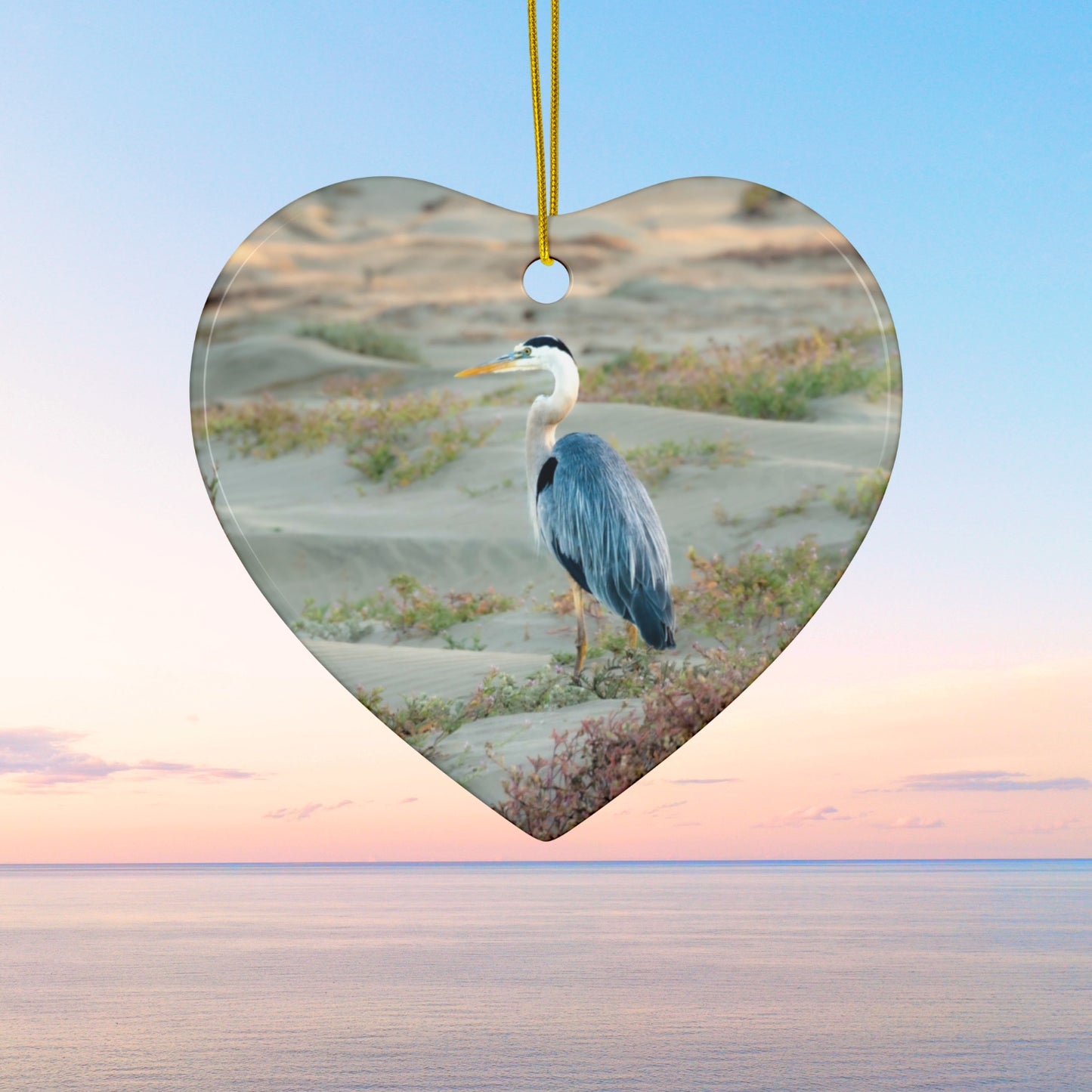Heart-shaped ceramic holiday ornament featuring a professional photograph of a Great Blue Heron standing among coastal reeds along the Pacific shoreline.