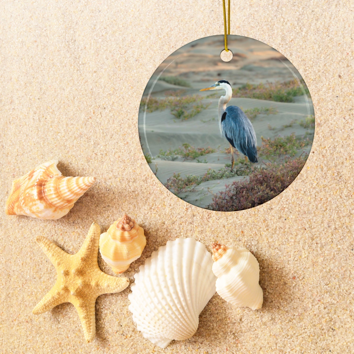 Ceramic holiday ornament featuring a professional photograph of a Great Blue Heron standing among coastal reeds along the Pacific shoreline on sand, surrounded by shells and a starfish.