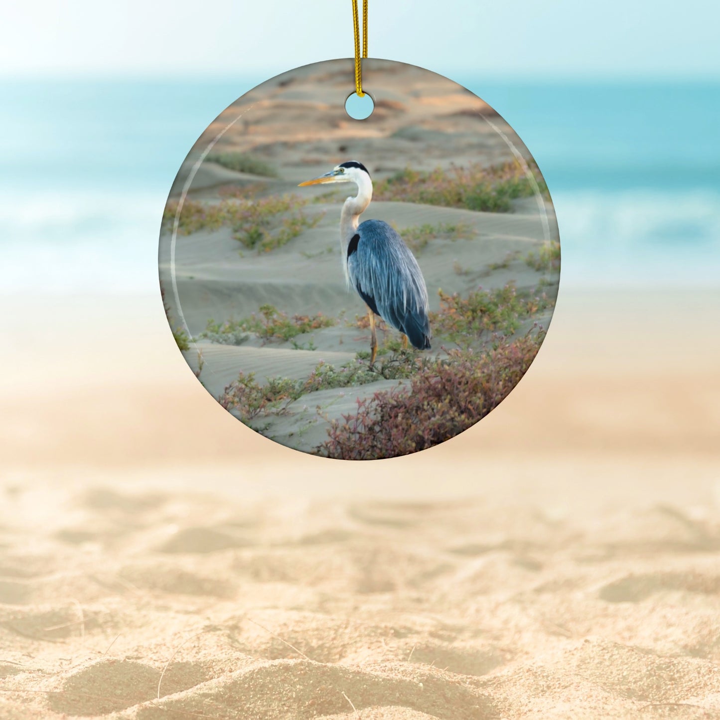 Ceramic holiday ornament featuring a professional photograph of a Great Blue Heron standing among coastal reeds along the Pacific shoreline.