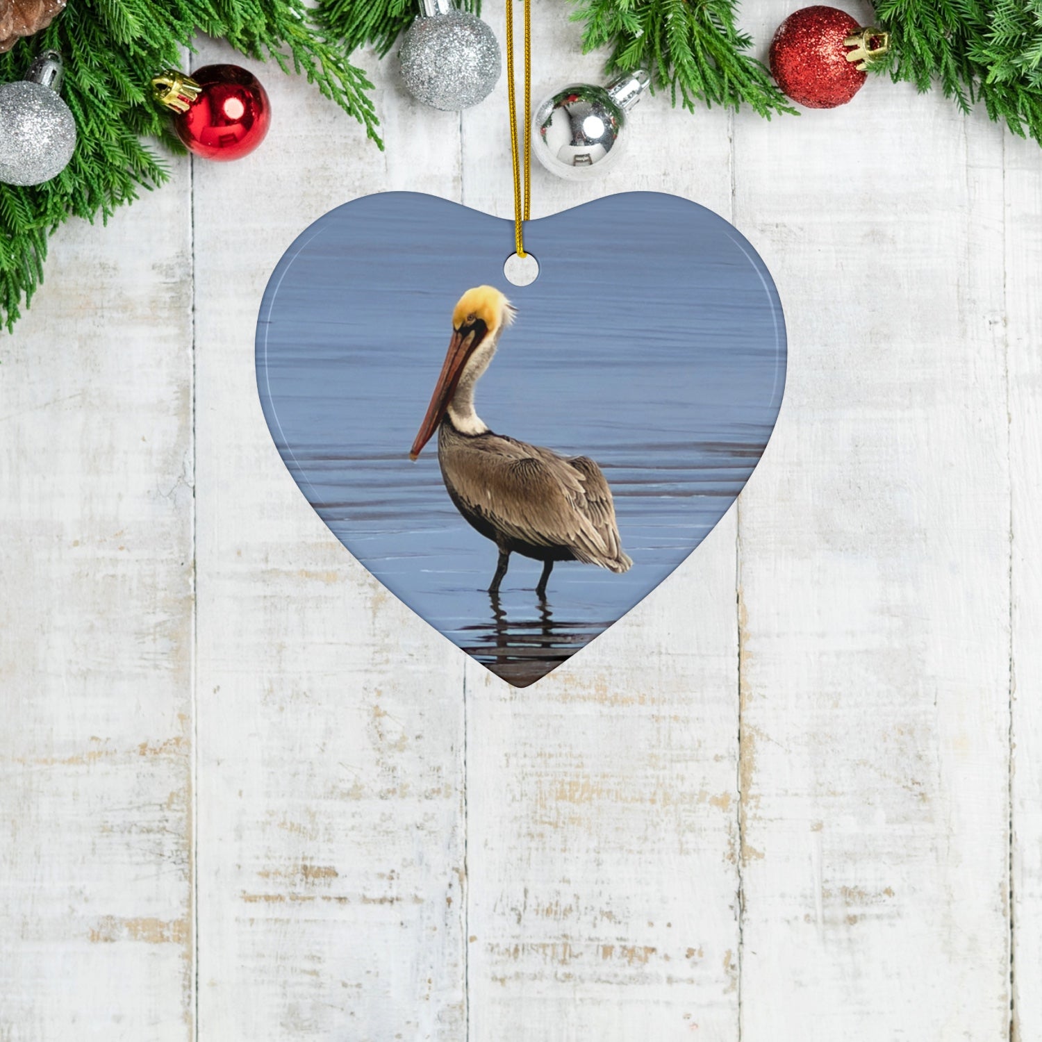 Heart-shaped ceramic holiday ornament featuring a professional photograph of a Brown Pelican standing on the shoreline  on a wooden surface with Christmas decorations.