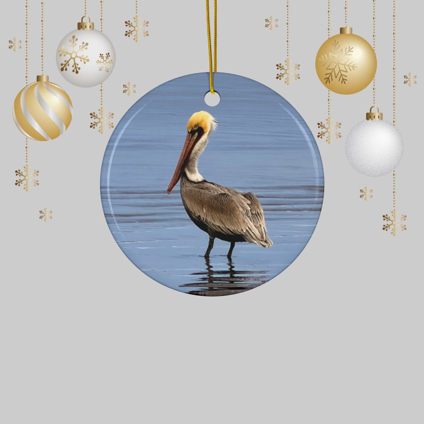 Ceramic holiday ornament featuring a professional photograph of a Brown Pelican standing on the shoreline on a gray background with Christmas ornaments.