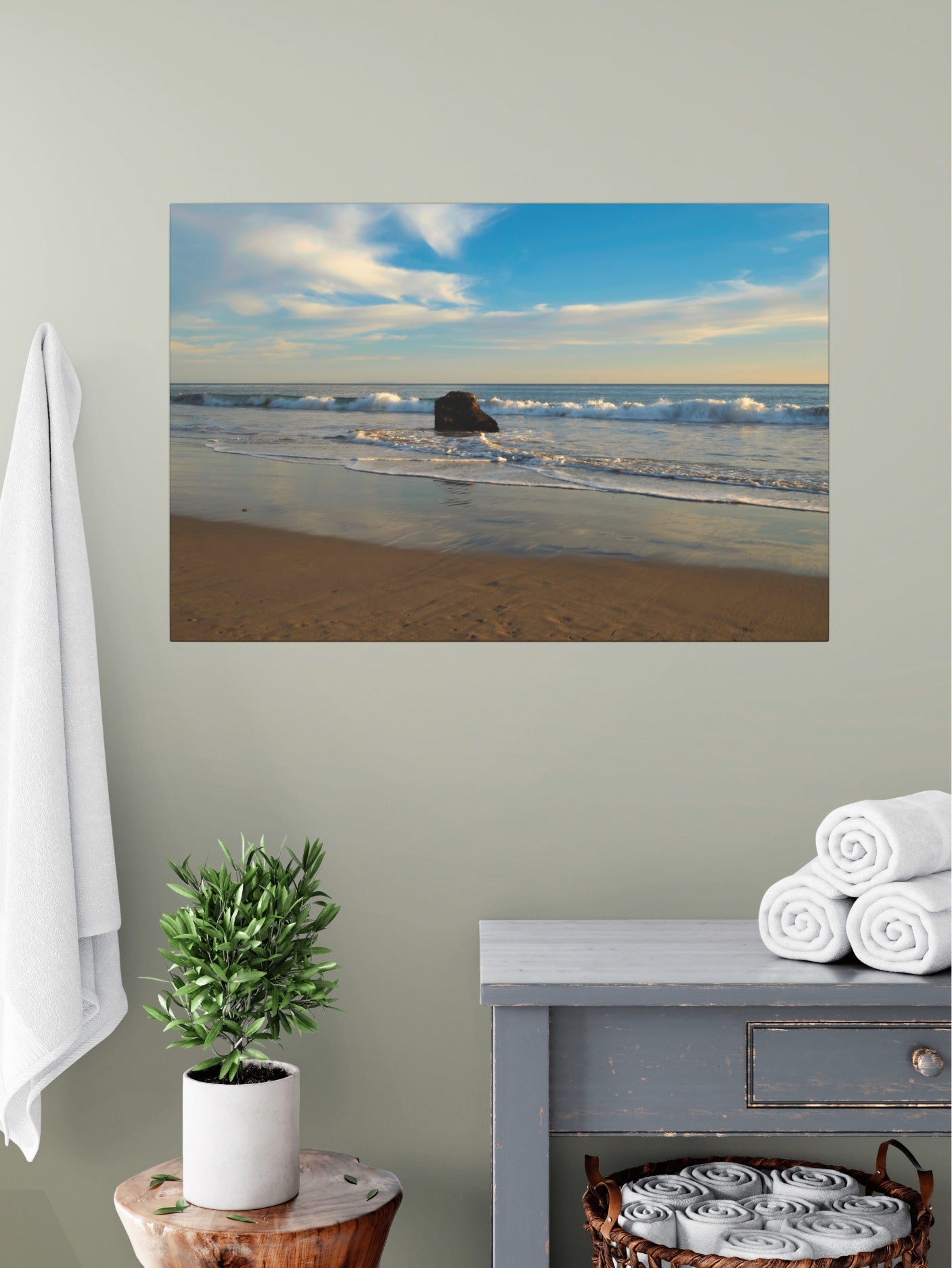 Solitary rock on a California beach with ocean wave foam and sky reflection canvas print on a wall above a table with a plant and folded towels.