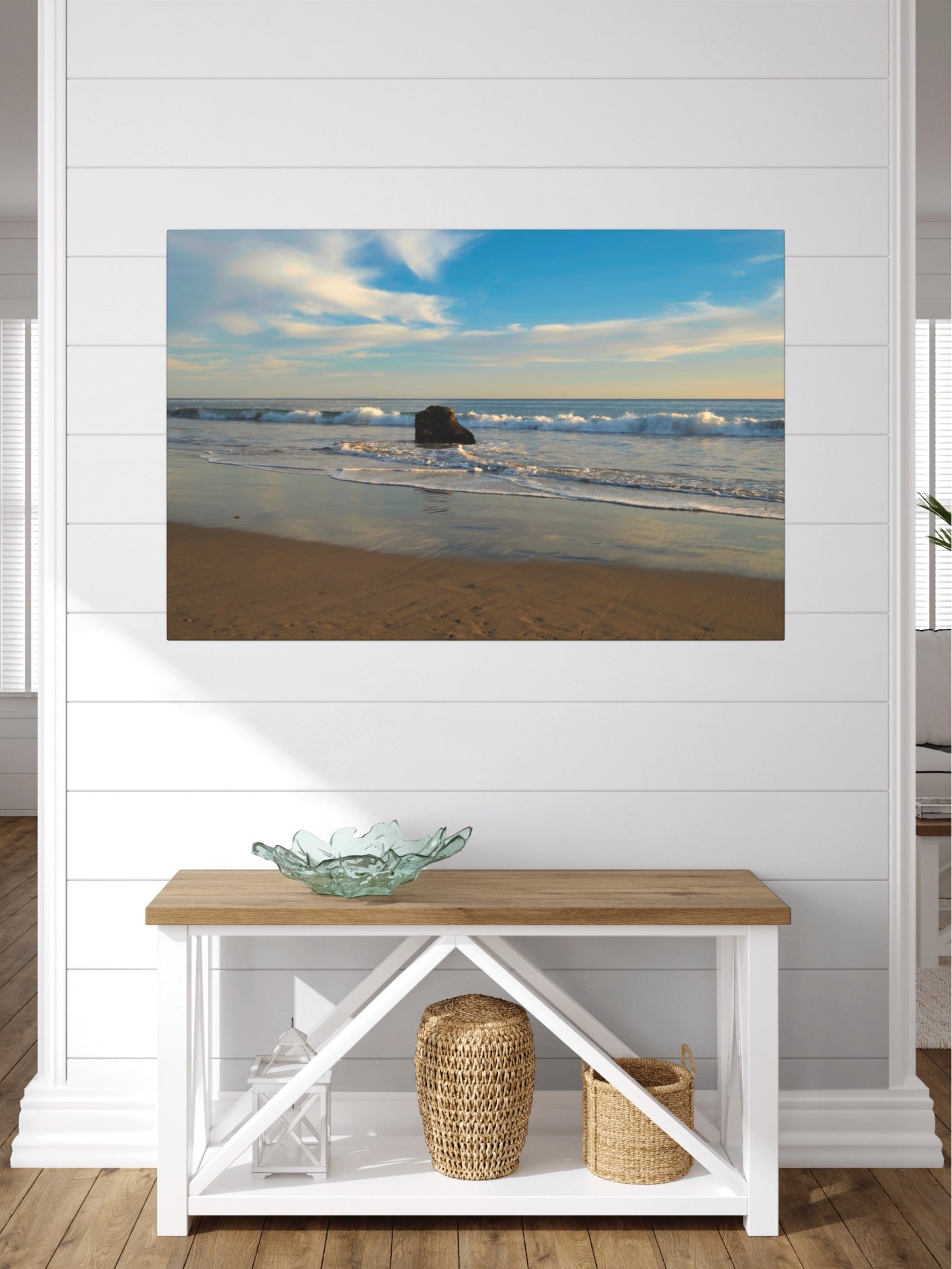 Solitary rock on a California beach with ocean wave foam and sky reflection canvas print on a wall above a wooden bench with storage baskets.
