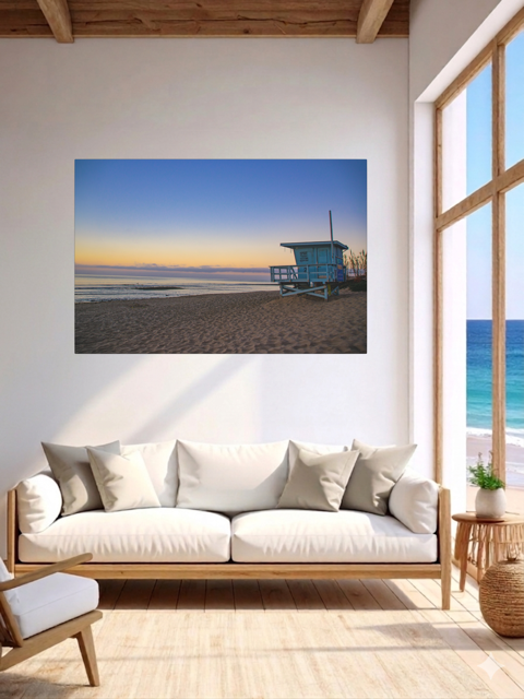 Living room with a canvas print of a California lifeguard tower at sunrise on the wall, white sofa, and large window.
