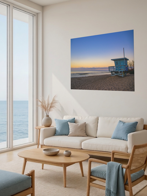 Living room with a large window view of the ocean, a beige sofa with blue cushions, and a canvas print of a California lifeguard tower at sunrise.