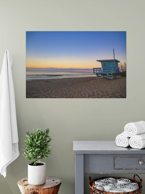 Canvas print of a California lifeguard tower at sunrise on a wall above a table with a plant and folded towels.