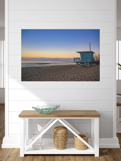 Canvas print of a California lifeguard tower at sunrise on a wall above a wooden bench with storage baskets.